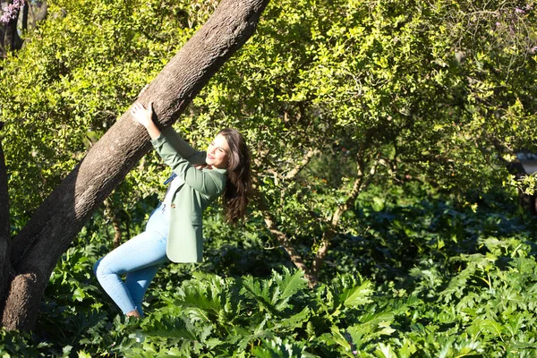 公園では 長い茶色の髪をした若い美しい女性がジーンズと緑のジャケットを手で木からぶら下げています 女性は幸せで楽しいです コンセプト幸福 — ストック写真