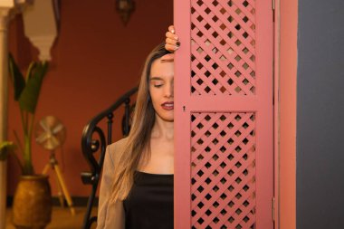 Beautiful young woman behind a pink wooden window cover. The woman is sad and looks at the ground with sorrow and depression.