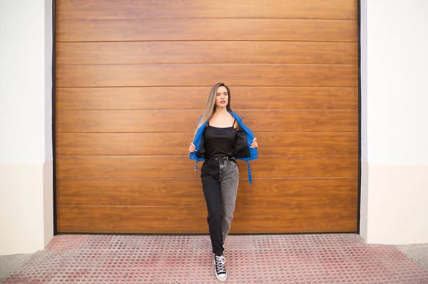 young and beautiful blonde woman dressed in black t-shirt and blue leather jacket and jeans on wooden background poses for photos doing different poses like a model. Beauty and fashion concept.