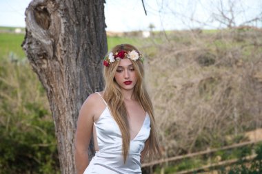 Uzun saçlı, beyaz elbiseli ve saçında çiçeklerden bir taç olan güzel sarışın kadın. Kadın çayırda, yeşilliğin ortasında.