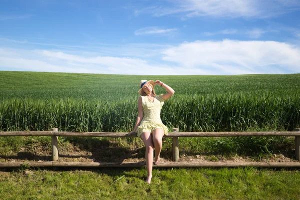 Blonde, young, pretty farm woman, sitting on a wooden fence. In the background a green wheat field. The woman is wearing a short green dress and a straw hat, which she is clutching in her hand