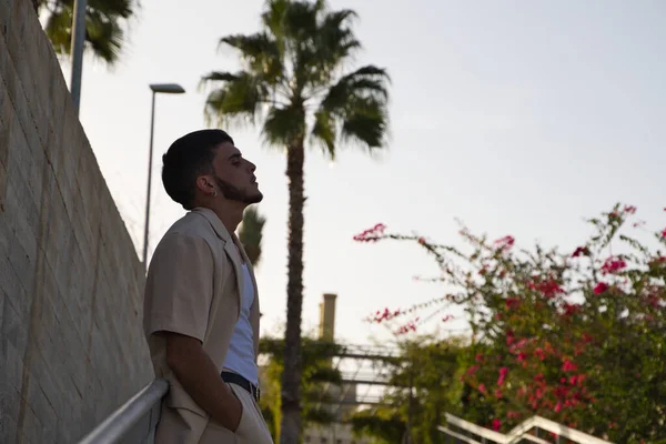 stock image Young Latin man dressed in beige suit, white t-shirt and black boots leaning on the railing of some park stairs. Man has social problems and is stressed thinking about life's difficulties.