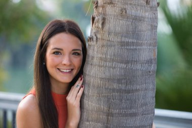 Young and beautiful woman leaning on the trunk of a palm tree. Woman is happy and relaxed and enjoying the sunny day in seville, spain. The woman is wearing jeans and orange top.