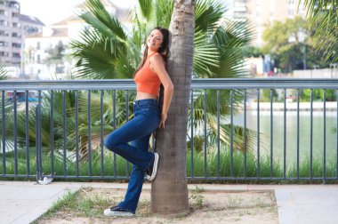 Young and beautiful woman leaning on the trunk of a palm tree. Woman is happy and relaxed and enjoying the sunny day in seville, spain. The woman is wearing jeans and orange top.