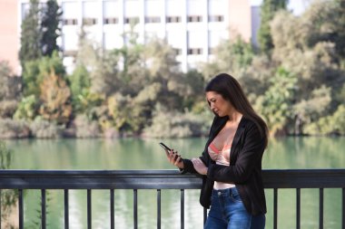 Young beautiful woman looking at her mobile phone leaning on the railing of the river in Seville, Spain. In the background you can see the river and part of the city.