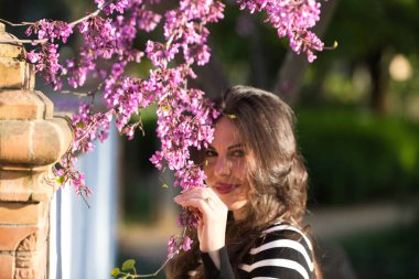 Uzun kahverengi saçlı, siyah beyaz çizgili gömlek giyen, pembe çiçekli bir ağaç dalının arasında saklanan güzel bir genç kadının portresi.