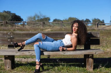 İspanyol, esmer, kıvırcık saçlı kadın ormana giden yolda bir bankta yatıyor. Kot pantolon ve beyaz bir bluz giyiyor ve güneşli günün tadını çıkarıyor..