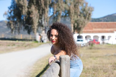 Kıvırcık saçlı esmer kadın kameraya bakarken kolunu patikanın ahşap çitlerinin üzerinden uzatıyor. Kot pantolon ve beyaz bir bluzla güneşli günün tadını çıkarıyor..