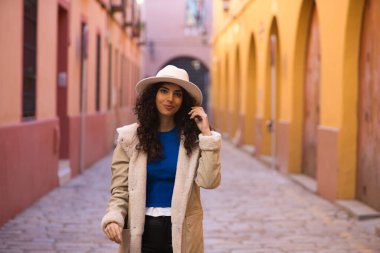 Young and beautiful Hispanic brunette woman with curly hair wearing a hat and coat for the cold walking in the city of seville while making different expressions and having fun.