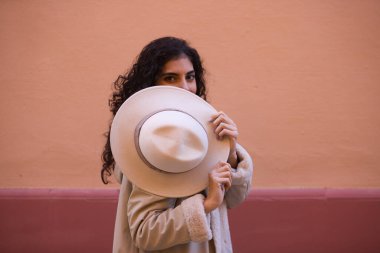 Young and beautiful Hispanic brunette woman with curly hair covers her face with her hat and exposes her eyes. The woman plays at hiding behind the hat making different funny expressions.