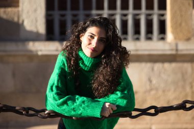 Kıvırcık saçlı ve yeşil yün ceketli genç, güzel esmer kadın İspanya 'daki Sevilla katedralinin etrafındaki zincirlere yaslanıyor. Kadın mutlu ve şehirde tatilinin tadını çıkarıyor..