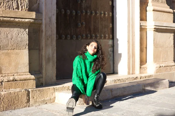巻き毛と緑のウールのコートを着たかなり若いブルネットの女性は スペインのセビリアの建物の入り口の一歩に座っています 女性は幸せと街で彼女の休日を楽しんでいます — ストック写真