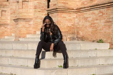 young and beautiful black latin woman wearing black clothes and sunglasses is sitting on the steps of the most important square in the city of seville, spain. The photo is taken from the front.