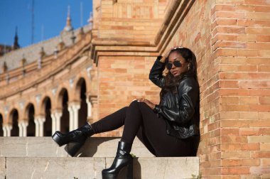 young and beautiful black latin woman wearing black clothes and sunglasses is sitting on the steps of a square in seville, spain. The photo is taken in profile and the girl is doing different poses.