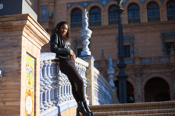молодая и красивая черная латиноамериканка находится в отпуске и посещает самую важную площадь города Севилья. Женщина одета в черное и опирается на перила знаменитой площади.