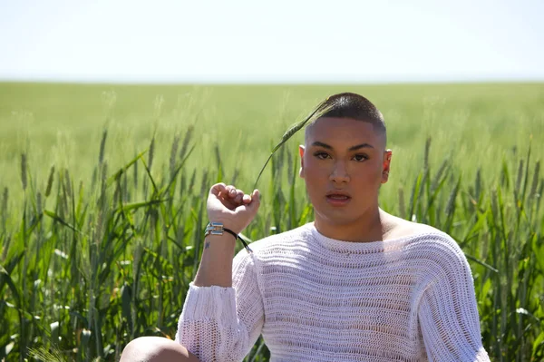 A young non-binary latin person is sitting in the wheat field and holds a green ear of wheat in his hand. In the background the wheat is growing and the sky is blue. Concept of organic agriculture.