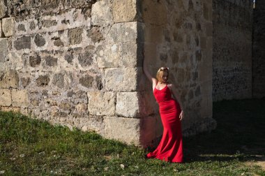 Sarışın, uzun saçlı, genç ve güzel, uzun kırmızı bir elbise giymiş ve başında çiçeklerden bir taç, bir kalenin duvarına yaslanmış. Kadın farklı ifadeler kullanıyor.