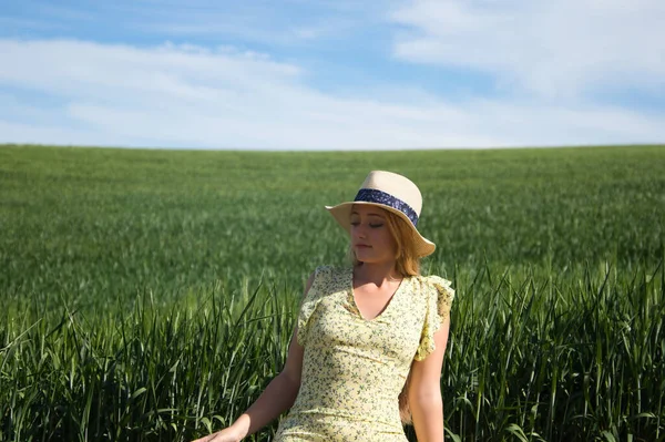Blonde, young, pretty farm woman, sitting on a wooden fence. In the background a green wheat field. The woman is wearing a short green dress and a straw hat and is happy and smiling