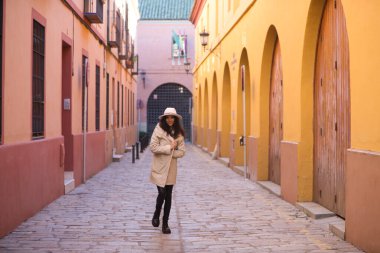 Young and beautiful Hispanic brunette woman with curly hair wearing a hat and coat for the cold walking in the city of seville while making different expressions and having fun.