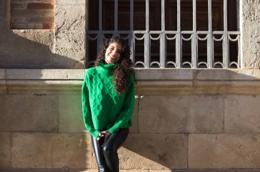 Genç ve güzel İspanyol kadın, kıvırcık saçlı ve yeşil ceketli esmer duvara yaslanıyor. Kadın mutlu ve Seville 'de güneşli bir günün tadını çıkarıyor..