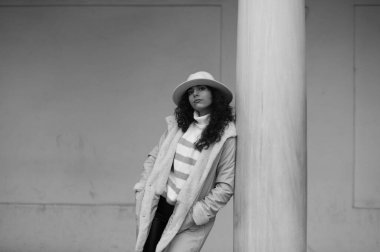 Genç ve güzel esmer, kıvırcık saçlı, bej şapkalı ve yünlü kışlık ceketli mermer bir sütuna yaslanmış bir kadın. Fotoğraf siyah beyaz çekilmiştir..