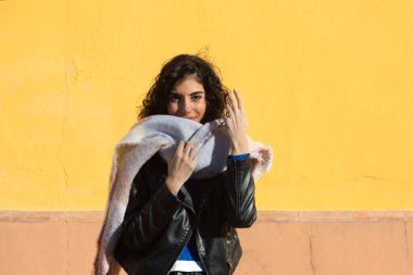 pretty young woman with curly brunette hair against a yellow background is dressed in winter clothes and wearing a scarf to protect herself from the cold. The woman is happy and having fun.