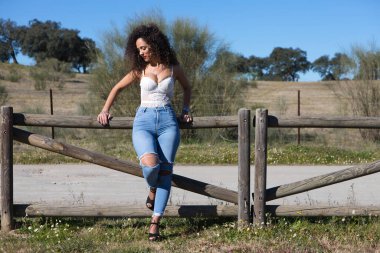 Kıvırcık saçlı, esmer, İspanyol kadın çitlerin üzerinde dinleniyor, çayıra gidiyor. O, kot pantolon ve beyaz bir bluz giydi ve güneşli günün tadını çıkarıyor. Kadın üzgün ve depresif..