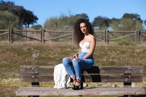 スペイン人 ブルネット 巻き毛の女性が森につながる道のベンチに座っています 彼女はジーンズと白い上に身を包み 晴れた日を楽しんでいます — ストック写真