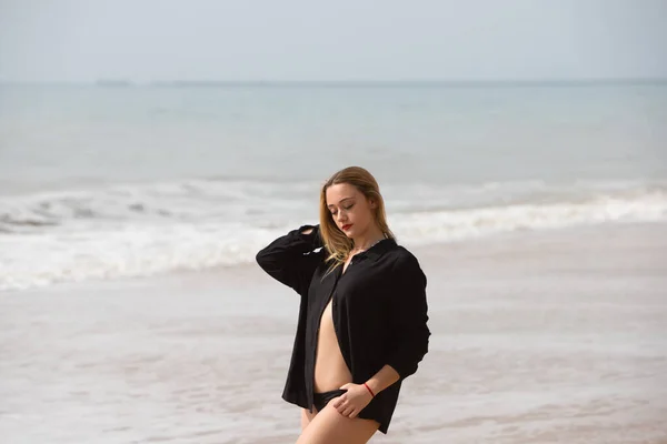若くて美しいブロンドの女性は黒のシャツと黒のビキニを着たビーチの海岸に沿って歩き 異なる表情や姿勢を作ります 海と空を背景に — ストック写真