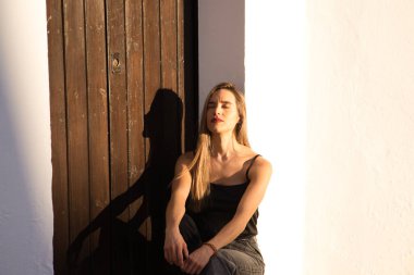 Young blonde woman dressed in black t-shirt and jeans sitting on the step of a Mediterranean style house in Spain. Woman closes her eyes and sunbathes.