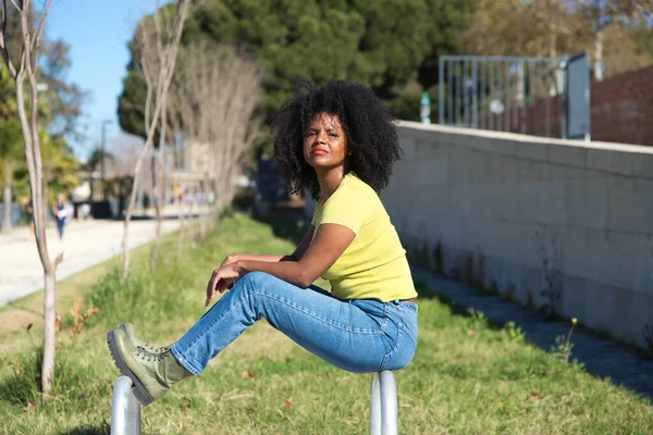 Young, pretty black woman with afro hair dressed in jeans and yellow shirt sitting in the bicycle car park of the park. The woman is sad and nostalgic.