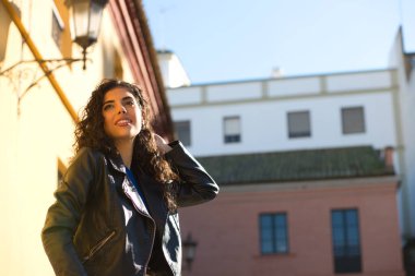 Young and beautiful Hispanic brunette woman with curly hair and black leather clothes is sitting on a railing in the street. The woman is happy and smiling.