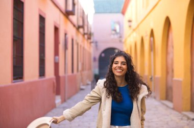 Young and beautiful Hispanic brunette woman with curly hair holds up a hat with her hand while making different expressions and having fun on her city trip in Europe.