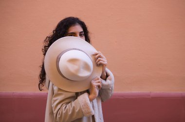 Young and beautiful Hispanic brunette woman with curly hair covers her face with her hat and exposes her eyes. The woman plays at hiding behind the hat making different funny expressions.