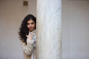 Young and beautiful brunette woman with curly hair and a woollen coat for the winter, leaning on a marble column while making different facial and body expressions.