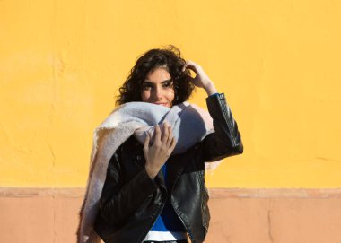 pretty young woman with curly brunette hair against a yellow background is dressed in winter clothes and wearing a scarf to protect herself from the cold. The woman is happy and having fun.