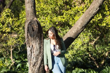 Uzun kahverengi saçlı, kot pantolon ve yeşil ceketli, İspanya 'da Seville' de bir parkta ağaca yaslanmış genç, güzel İspanyol bir kadın. Kadın dalgın ve yere bakıyor.