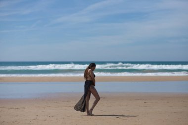 Yüzme kostümlü, uzun kahverengi saçlı ve kumsalda siyah ipekli sarong giyen güzel genç bir kadın güneşli günün huzur ve sessizliğinin tadını çıkarıyor. Arka planda mavi deniz
