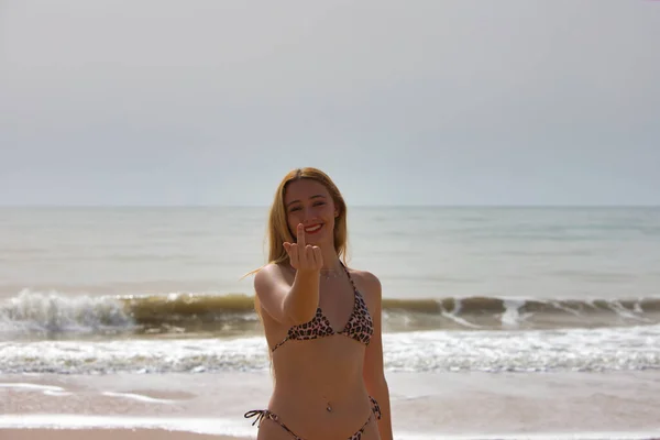 かなり若いブロンドの女性が不満を示すために中指を上げます 女性はビーチの岸にいて幸せ — ストック写真