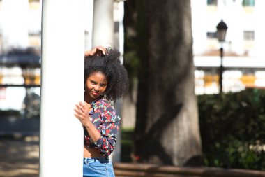Parktaki mermer bir sütuna yaslanmış, afro saçlı, genç, güzel siyahi bir kadın. Kadın farklı ifadeler kullanırken güneşli bir günün tadını çıkarıyor.