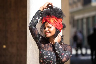 Genç, güzel, siyah bir kadının portresi, afro saçlı, çiçekli siyah elbiseli, saçında kırmızı bir eşarp olan. Kadın mutlu, gülüyor ve mermer bir sütuna yaslanarak eğleniyor.