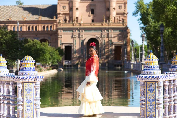 stock image Young black woman dressed as a flamenco gypsy in a famous square in Seville, Spain. She is wearing a beige dress with ruffles and a red shawl and is in front of a canal in the square