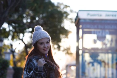 Ekose ceketli, yün şapkalı ve güneş gözlüklü güzel bir kadın parkta güneşli kış gününün tadını çıkarıyor. Kadın mutlu ve gülümsüyor..