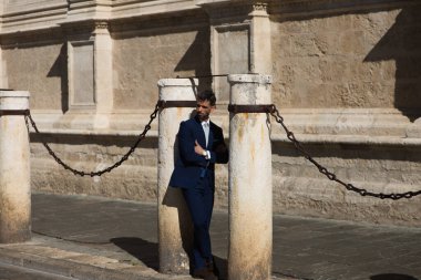 Sakallı, mavi takım elbiseli ve kravatlı yakışıklı bir genç kollarını İspanya 'daki Sevilla Katedrali' nin koluna yaslıyor. Bu adam şehirde bir iş gezisinde olan bir yönetici.