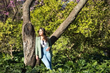 Uzun kahverengi saçlı, kot pantolon ve yeşil ceketli, İspanya 'da Seville' de bir parkta ağaca yaslanmış genç, güzel İspanyol bir kadın. Kadın düşünceli ve üzgün ve yana bakıyor.