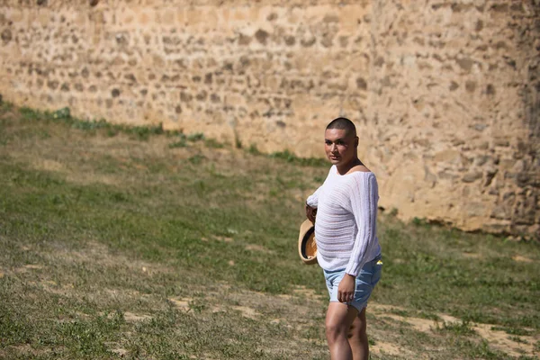Portrait of young non-binary latin person walking with a straw hat in his hand while having fun. In the background a medieval castle. Concept of diversity, homosexuality and human rights