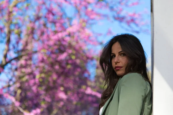 stock image Portrait of a beautiful young woman with long brown hair and a serious and sad character. The woman is looking at the camera. In the background pink flowers and blue sky. Concept of depression.