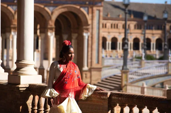 Young black woman dressed as a flamenco gypsy in a famous square in Seville, Spain. She wears a beige dress with ruffles and a red shawl with flowers. Flamenco cultural heritage of humanity