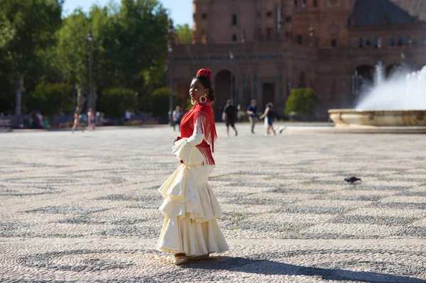 Young black woman dressed as a flamenco gypsy in a square in Seville, Spain. She wears a beige dress with ruffles and a red shawl with flowers. Flamenco cultural heritage of humanity. Dance concept
