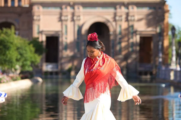 Young black woman dressed as a flamenco gypsy in a famous square in Seville, Spain. She is wearing a beige dress with ruffles and a red shawl and is in front of a canal in the square